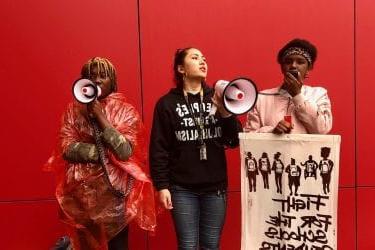 三名妇女站在红墙前，举着标语和扩音器